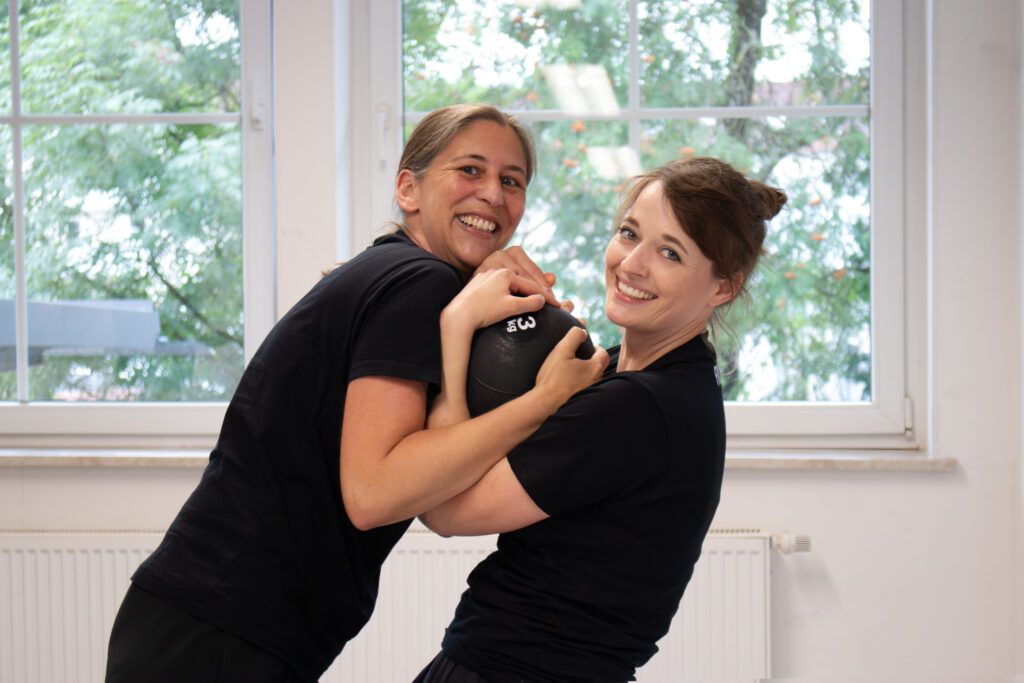 Zwei Frauen kämpfen um einen Medizinball und haben dabei sichtlich Spaß.