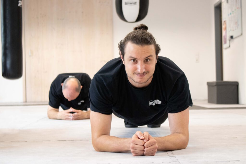 Ein Mann macht im Kampfsporttraining die Plankübung und lächelt.