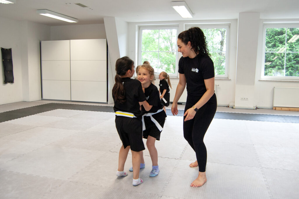 Zwei Kinder aus dem Selbstverteidigungskurs üben eine Griffbefreiung ein während die Trainerin Hilfestellung gibt.