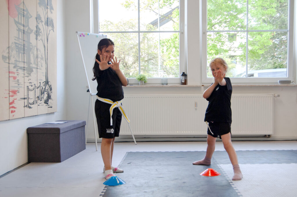 Zwei Kinder aus dem Selbstverteidigungskurs stehen vor einem Kegel und nehmen eine Abwehrhaltung ein.