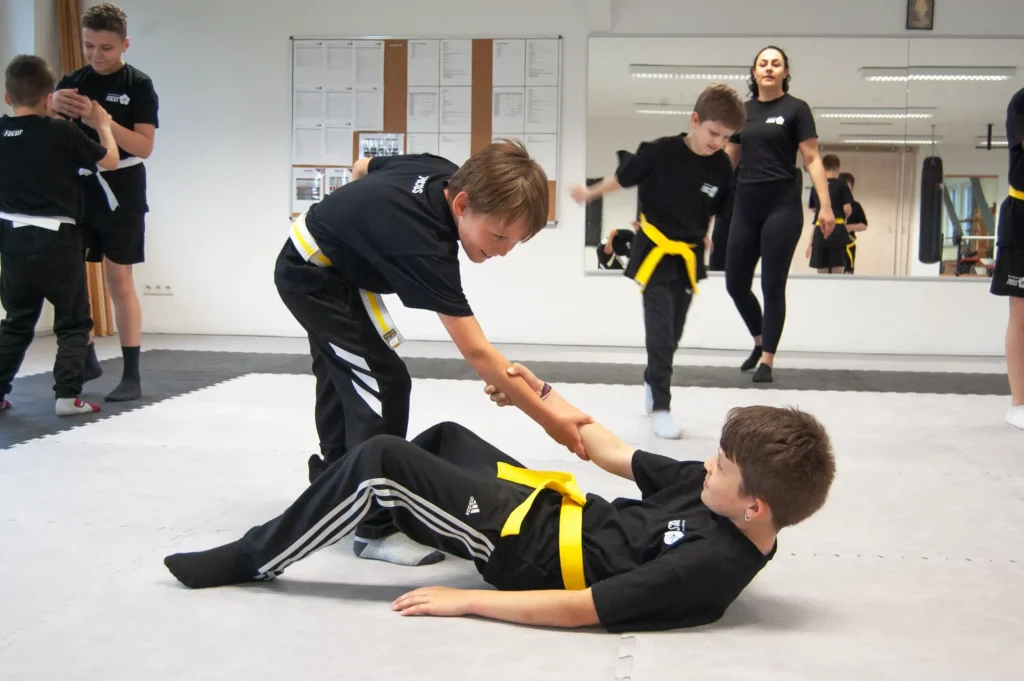 Ein Kind hilft einem anderen Kind vom Boden aufzustehen. Kinder lernen wichtige Werte in der Kampfsportschule.